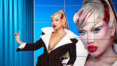 A blonde drag queen in a business-style suit and high ponytail poses in front of an image of herself