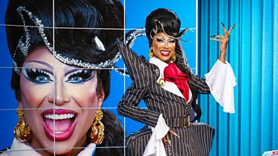 A drag queen (Serena Morena) poses in a pinstripe suit with high shoulder pads and a bejeweled wig. 