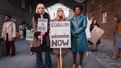 Three midwives stand together in their 70s style civilian clothing at a protest holding signs that say 'Venus is Here' and 'Equal Pay Now'