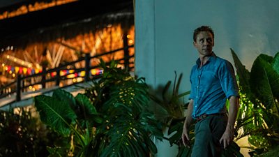 A man in a shirt and trousers lurks in the bushes outside a restaurant looking shocked