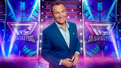 A man in a blue suit smiles to camera, set against the backdrop of the Gladiators arena