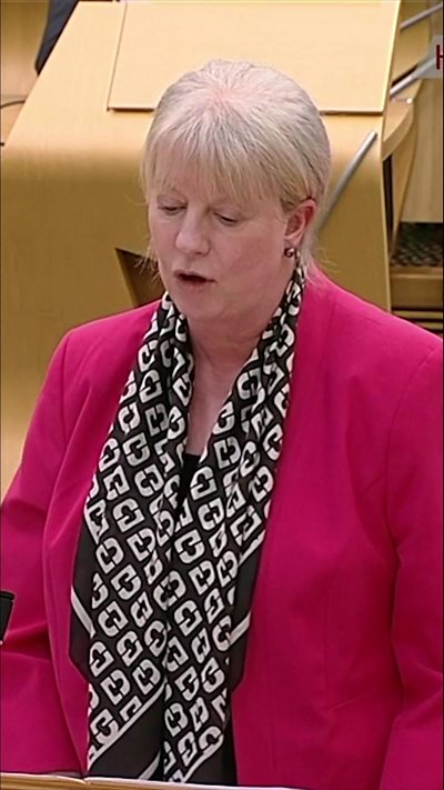 Woman with short blonde hair in a pink jacket and black and white scarf.