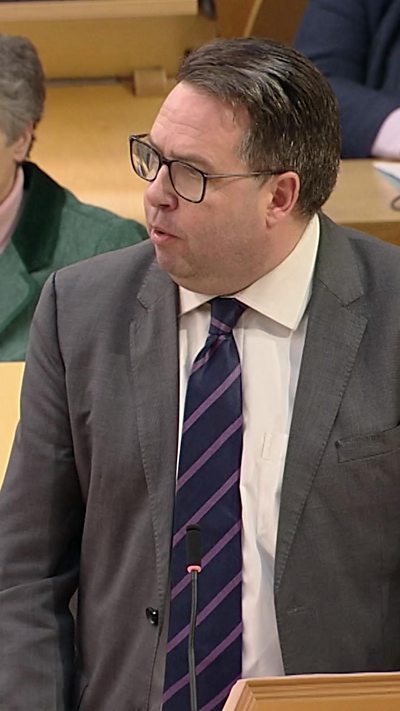 Man with short dark hair and glasses in grey suit and dark tie.