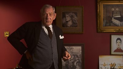 Ian McKellen in character as L.S. Lowry. He stands in a suit, tie and cardigan beside a wall covered in artwork. 