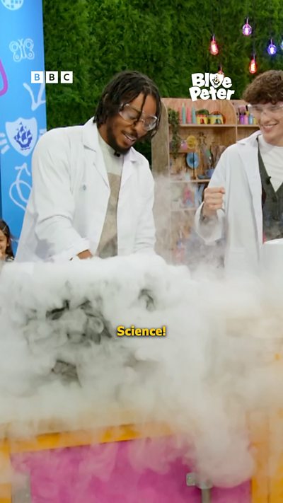 Big Manny and Joel wearing lab coats and safety goggles whilst a cauldron bubbles over and emits lots of fog.