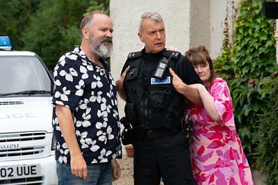 A man in a floral shirt stands by a police car as a woman hangs on to a police officer