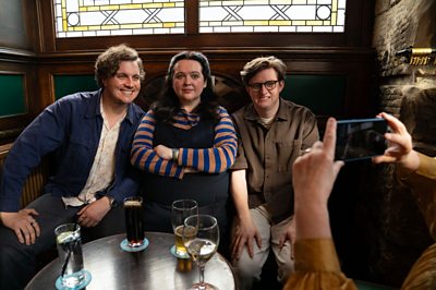 Two men and one woman pose for a photo in a pub - the characters are Clay (Hyoie O' Grady), Nina (Ashley Storrie), Lee (Lorn Macdonald) posing for Dinosaur