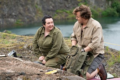A woman and a man on an excavation site smile at each other as they examine the ground