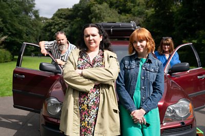 Nina (Ashley Storrie) and Evie (Kat Ronney) stand in front of a car in a still for Dinosaur, Series 2