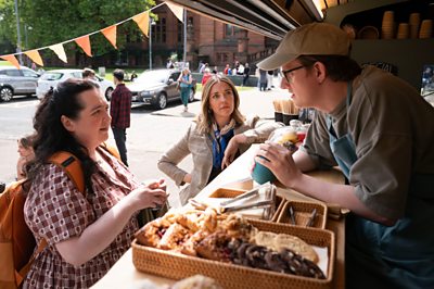 A woman - Nina (Ashley Storrie) talks to a man at a baked goods stand while another woman looks on