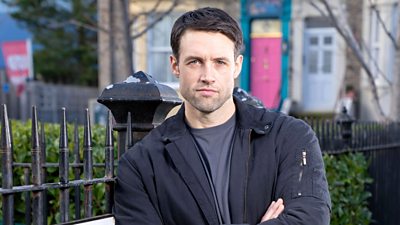 A man in a black jacket and a grey t-shirt stands with his arms folded, leaning against the railings in Albert Square