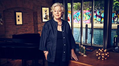 A woman in a dark denim shirt and black top stands by a desk, looking to camera