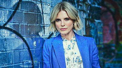 A woman in a flowery blouse and blue blazer leans against a wall that's been covered in graffiti