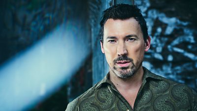 A man in a green patterned shirt looks to camera as a light shines through the darkness behind him