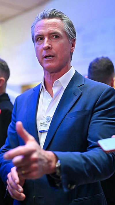 Gavin Newsom wears a blue suit, white shirt and no tie while speaking to reporters.