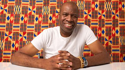 A man in a white t-shirt smiles as he sits at a table, set against a patterned blue, orange and yellow background