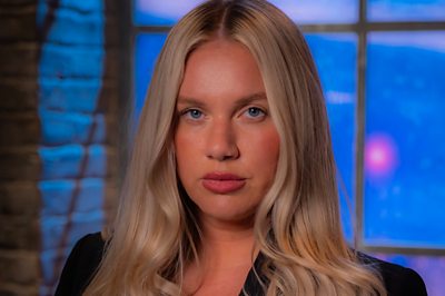 A blonde woman (Jenna Meek) stands in a dark mysterious room 