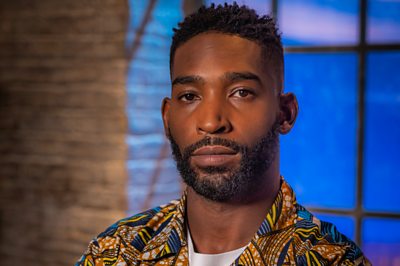 A man in a fun-printed shirt (Tinie Tempah) poses in a moody room with a dark window.