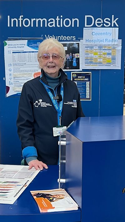 A woman with grey hair and glasses in a blue top at a reception desk