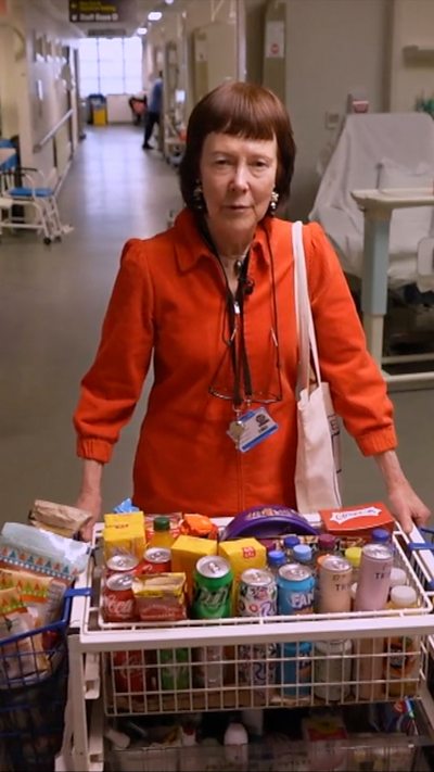 A person in a red outfit stands in a hospital corridor beside a cart filled with various snacks.