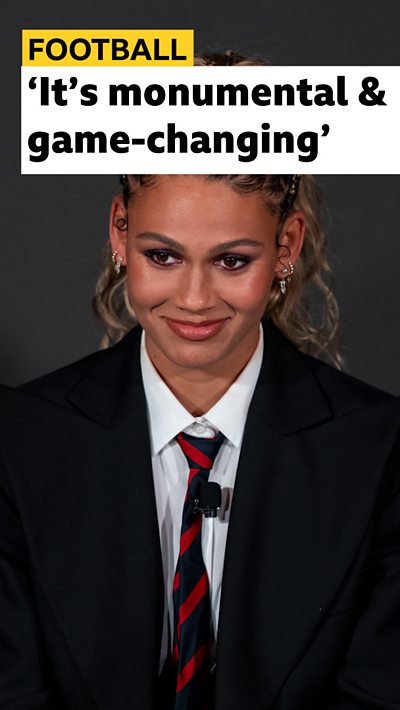 Washington Spirit forward Trinity Rodman smiling while signing her new contract. She is wearing a suit and tie.