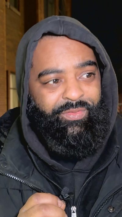 man wearing a hooded swaeatshort and coat with a large beard speaks into a small microphone.