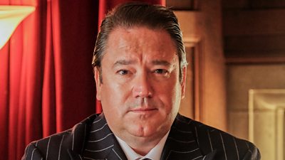 Un hombre con un traje de raya diplomática (el Sr. Tramley, de Boarders) mira a la cámara.