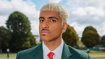 Un estudiante rubio (Yusuf, Boarders) con uniforme verde posa en un parque.