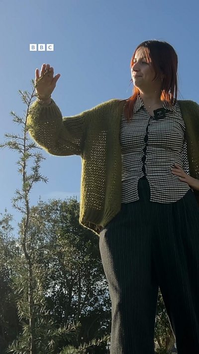 A woman with red hair stands next to a sapling