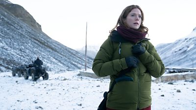 First look from Vigil series three. DI Kirsten Longacre (Rose Leslie) stands outdoors between snow-covered hills. Wearing warm winter clothing and carrying a rifle, she looks at something offscreen with concern. A figure on a quad bike is visible in the background. 