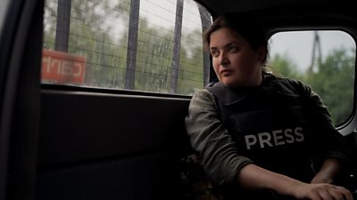 A woman in a bulletproof vest labelled 'press' sits in the back of a vehicle, looking out through the back window