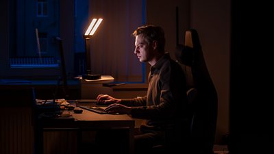A man leans over a laptop keyboard lit by a single lamp 
