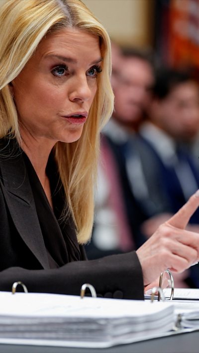 A woman with blonde hair in a dark suit pointing her index finger.