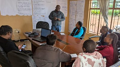 A group of radio journalists in Zambia are seated around a table listening to a �鶹�� Media Action mentor giving training about AI, the walls are yellow and there are notes and papers all over the walls behind him that the group have participated in creating. 