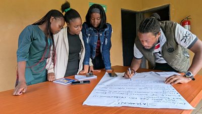 Four radio journalists are working together around a table, they have a big piece of white paper and are working together to develop a statement with a blue felt tip pen, a man is doing the writing, three women are inputing ideas, the walls behind them are yellow and they are in a radio station in Zambia.