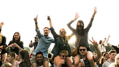 A scene from a festival crowd - looking into the crowd, three people are sat on other people's shoulders, enjoying the show.