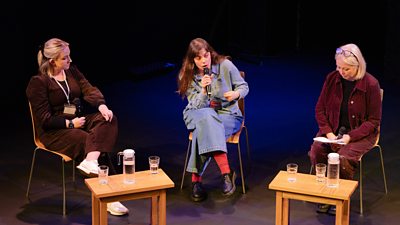 Three people sit on stage in chairs having a panel-style conversation, with small tables holding water glasses and bottles in front of them.