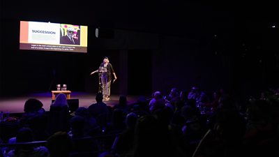 A person stands on stage giving a presentation in front of an audience, with a large screen displaying a slide behind them and a small table with water glasses nearby.