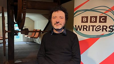 The writer Jeremy Dyson sat ready to be interviewed in front of a stand up banner which reads BBC Writers with a graphic design