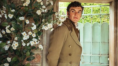 A man in formal clothing from the 18th Century leans against a stone doorway to a garden. White flowers grow alonside the wall beside him. 