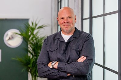 A man (Tom Kerridge) poses with his arms folded in a corridor.