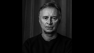 A black and white head and shoulders shot of a man in a dark jumper