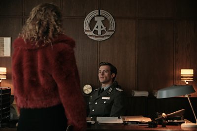 A woman approaches a man at his desk, dressed in army uniform.