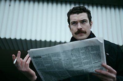 A man in a moustache looks at something off camera as he holds up a newspaper