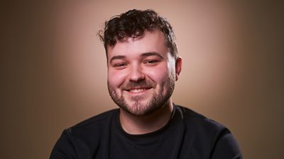 A man in a black t-shirt smiles to camera, set against a brown background