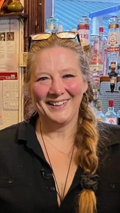 A woman with ginger hair in a pony tail wearing a black top stood behind a pub bar