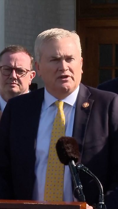 A man with grey hair in a dark suit and yellow tie stands at a podium