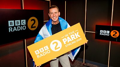 Scott Mills stands in the BBC Radio 2 studio wrapped in a Scottish flag holding a board that reads BBC Radio 2 in the Park Stirling 2026