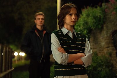 Two people stand in a dark outdoor setting. The main woman in the foreground has her arms folded looking thoughtful.