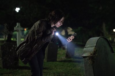 A young woman is in a graveyard with a torch and phone.